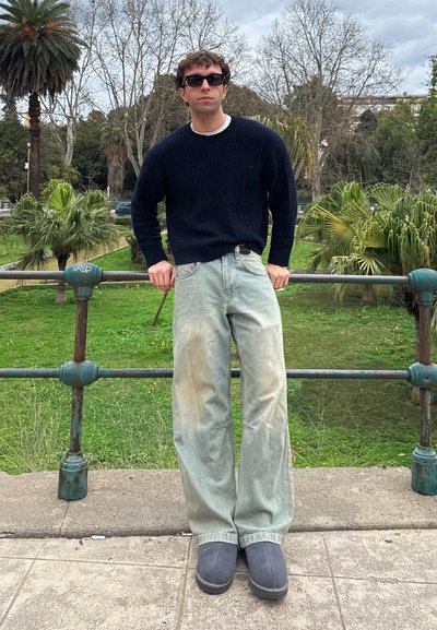 Hombre con suéter negro, pantalones anchos claros, zapatos grises y gafas de sol apoyado en una barandilla de metal en un parque con palmeras y cielo nublado.