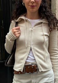 Beige zip-up hoodie med ribbet tekstur og snører, lagd over en hvid skjorte. Brun bælte med metalstuds, håndtaske med vævet design.