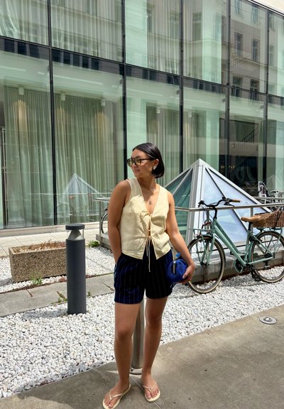 Top sin mangas con lazo frontal en color crema, pantalones cortos de rayas azules, gafas de sol y un bolso de mano azul. El fondo presenta un edificio de cristal y una bicicleta.