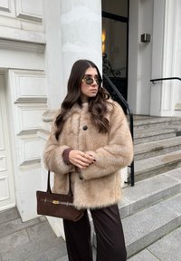 Femme aux longs cheveux noirs portant des lunettes de soleil, un manteau en fourrure beige, un pantalon foncé, tenant un sac à main marron, debout près des marches d'un bâtiment blanc.