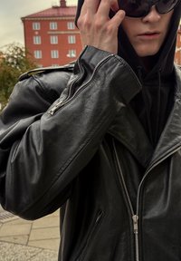 Schwarze Lederbikerjacke mit silbernen Reißverschlüssen; verfügt über einen breiten Reverskragen und Reißverschlusstaschen. Das Modell trägt einen schwarzen Hoodie darunter.