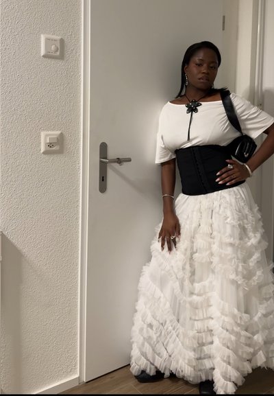 Mujer con falda blanca con volantes, top blanco con corsé negro, bolso de hombro negro y collar en forma de flor de pie junto a una puerta blanca cerrada.