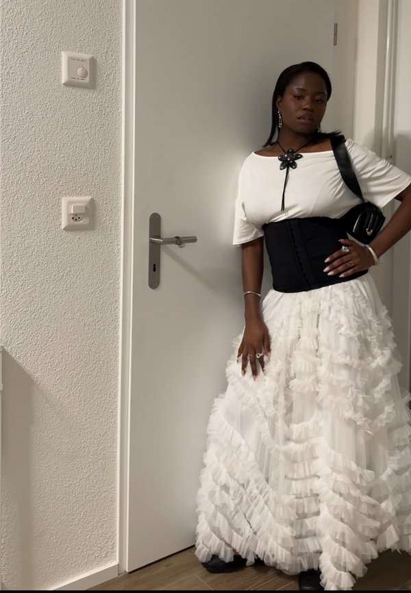Γυναίκα που φοράει λευκό φουσκωτό φόρεμα, λευκό τοπ με μαύρο κορσέ, μαύρη τσάντα ώμου και κολιέ σε σχήμα λουλουδιού, στέκεται δίπλα σε κλειστή λευκή πόρτα.