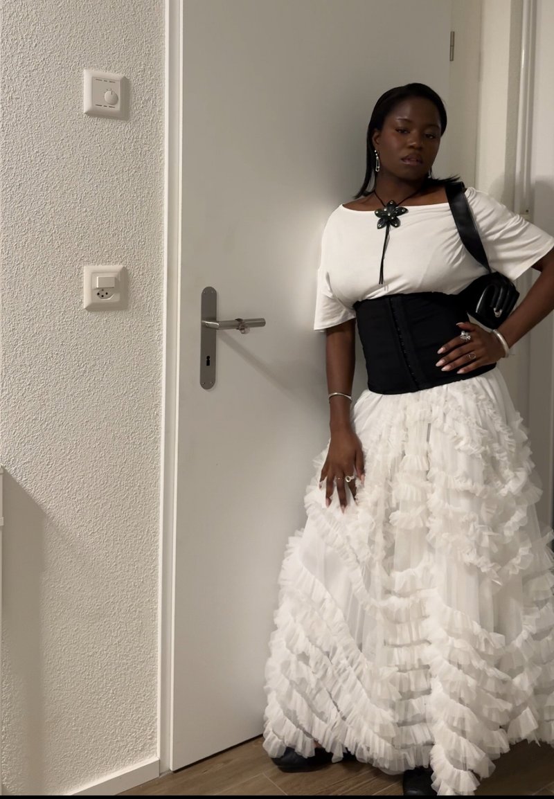 Kobieta w białej spódnicy z falbanami, białej bluzce z czarnym gorsetem, czarnej torbie na ramię i naszyjniku w kształcie kwiatu stoi przy zamkniętych białych drzwiach.