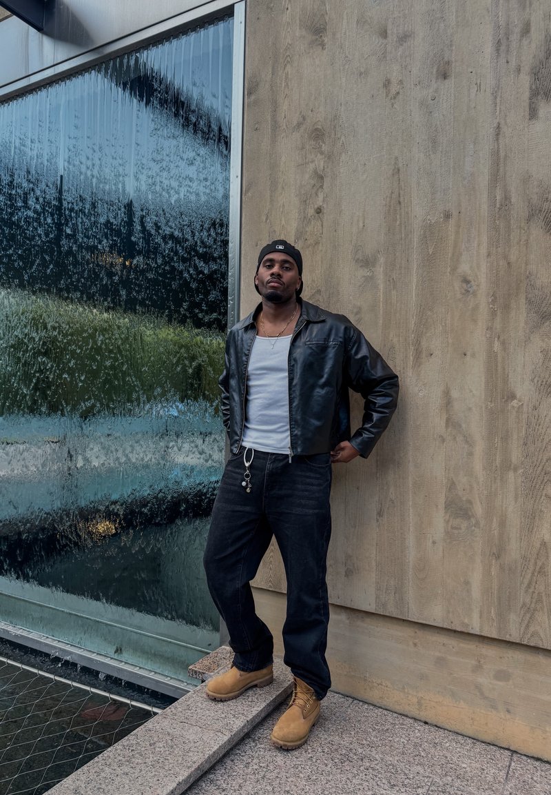 Black leather jacket, white vest, black jeans, tan boots. Standing against a textured wooden wall, near a water feature.