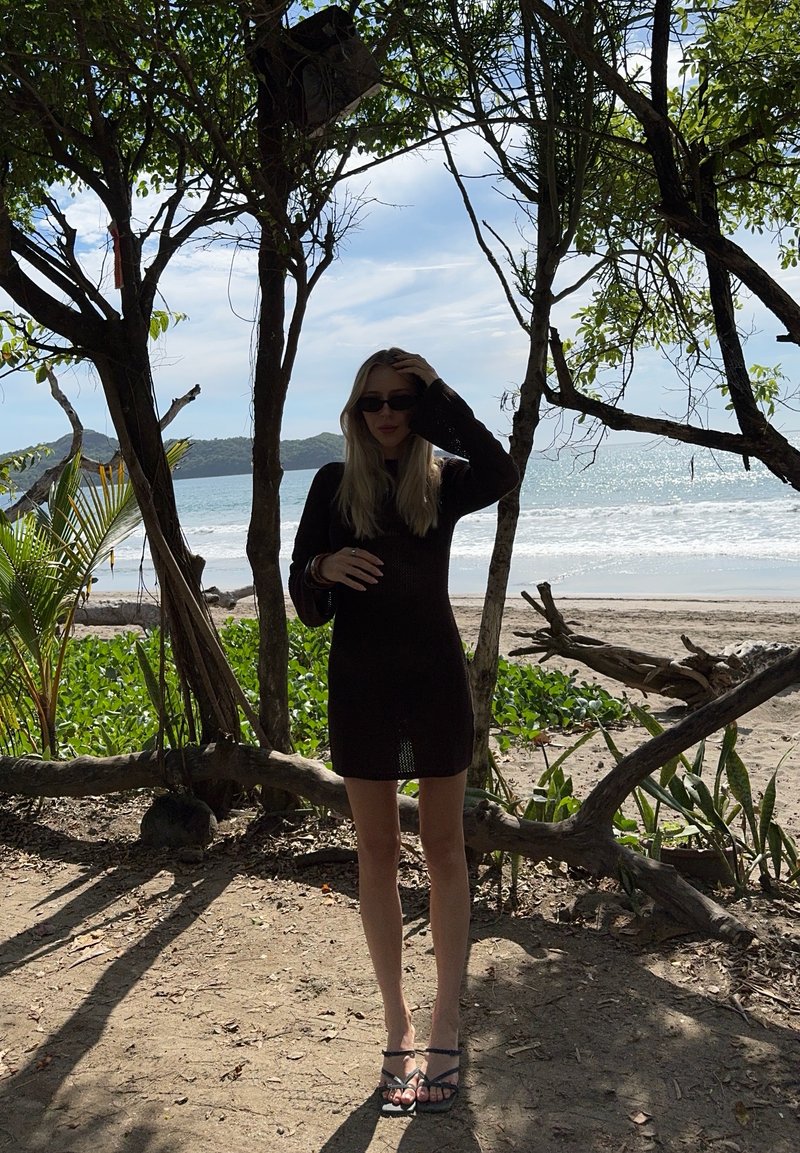 Frau in schwarzem Kleid und Sonnenbrille steht auf einem schattigen Strandweg, umgeben von Bäumen, mit Meereswellen und entfernten Hügeln im Hintergrund.