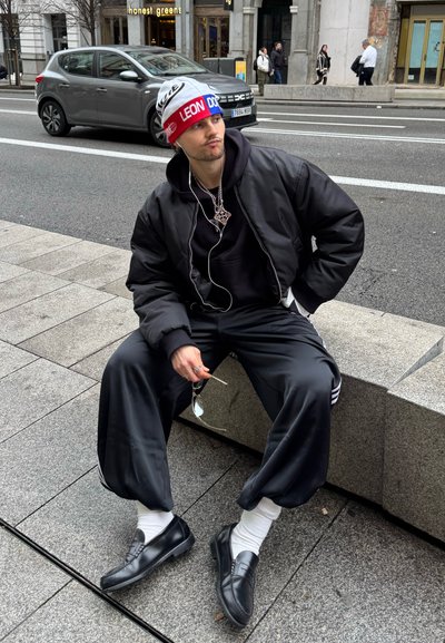 Joven con chaqueta negra, pantalones holgados y gorro colorido sentado en un banco de piedra cerca de una calle de la ciudad con coches y peatones.