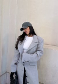 Grey wool overcoat with a tailored design, large lapels, and buttons. Worn over a white shirt, paired with a grey cap and a black handbag.