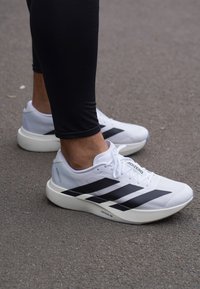 En person som bär vita Adidas Adizero löparskor med svarta ränder på asfalt, klädd i svarta leggings.
