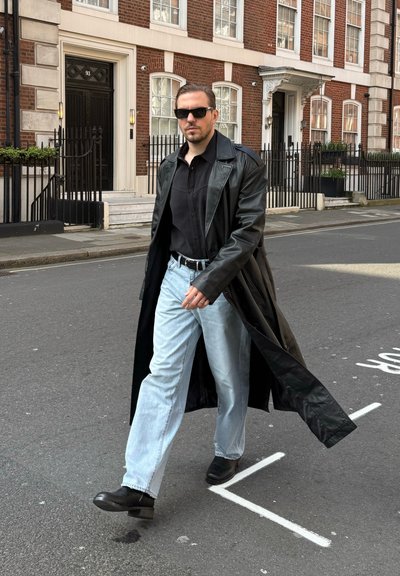 Hombre con abrigo largo de cuero negro, camisa negra, vaqueros azul claro, botas negras y gafas de sol caminando por una calle de ciudad con edificios de ladrillo.