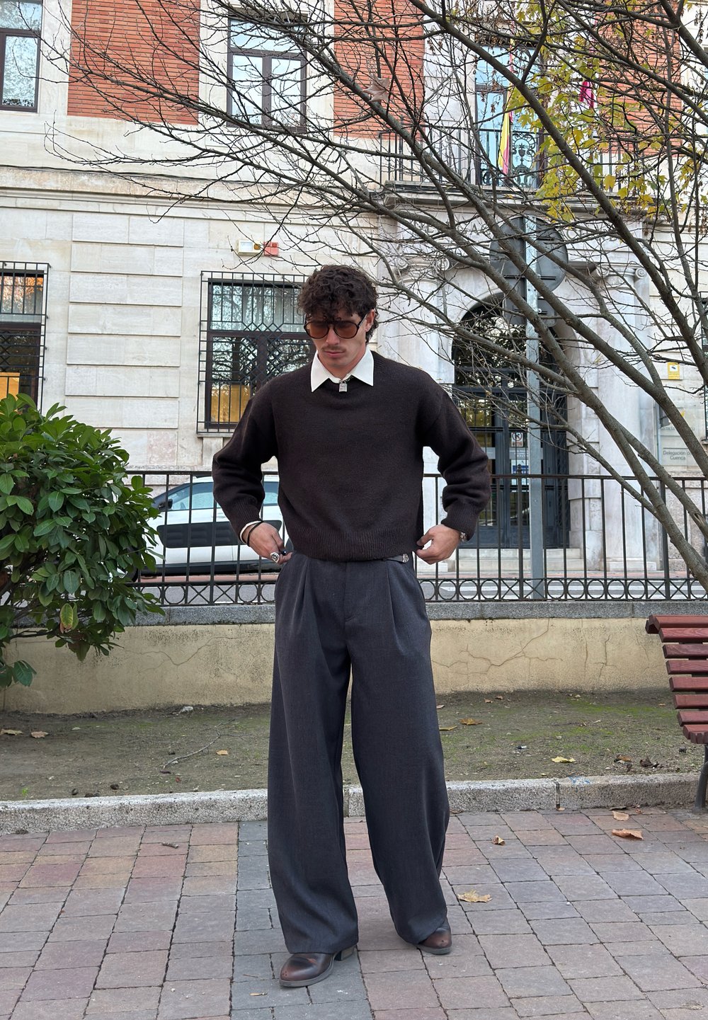 Tamsiai rudas megztinis, po juo baltas marškinių apykaklė, derinamas su laisvais tamsiai pilkais klostuotais kelnėmis ir rudos spalvos batais. Miesto lauko aplinka.