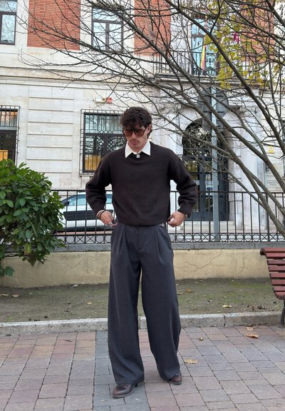 Jersey marrón oscuro con una camisa blanca de cuello debajo, combinado con pantalones plisados sueltos de color gris oscuro y zapatos marrones. Entorno urbano al aire libre.