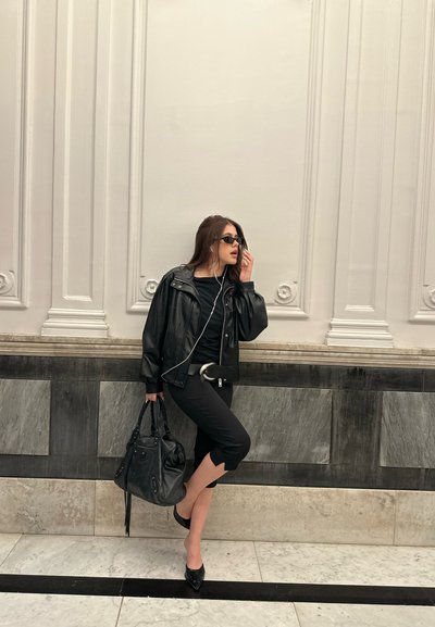Chaqueta de cuero negra, top y pantalones negros, tacones de punta negros, un gran bolso negro, gafas de sol y auriculares, contra una pared gris clara.
