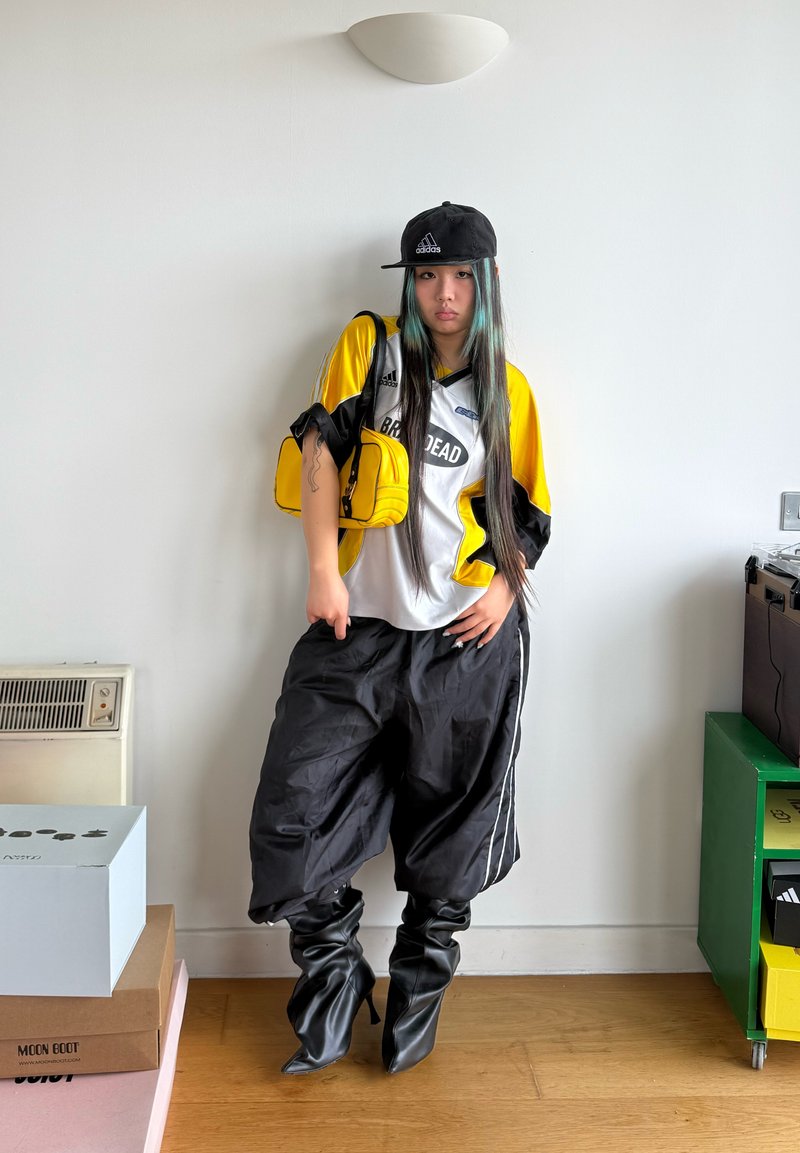 Jeune femme aux longs cheveux portant une casquette noire, un maillot de sport jaune et blanc, un pantalon noir ample et des bottines noires à talons, debout à l'intérieur.