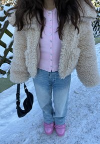 Personne portant une veste beige en peluche, un pull rose boutonné, un jean bleu clair et des chaussures roses, tenant un sac à main noir, debout sur un sol enneigé.