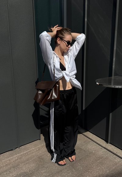 Camisa blanca de botones atada a la cintura, top de bikini negro, pantalones deportivos negros con rayas blancas, bolso de cuero marrón al hombro, gafas de sol.