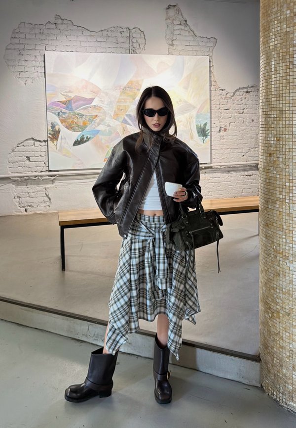 Femme portant des lunettes de soleil, une veste en cuir noire, un top court blanc, une jupe à carreaux et des bottes noires, tenant une tasse et un sac à main dans une galerie d'art.