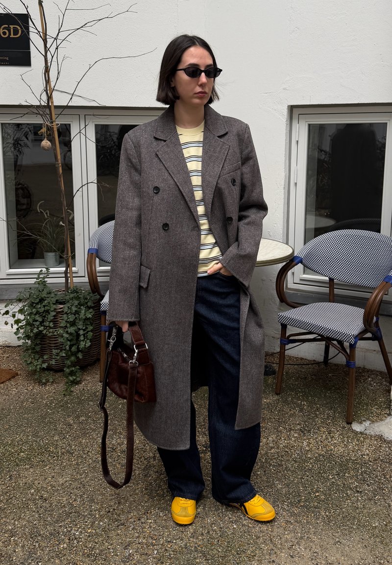Femme portant un long manteau gris, chemise à rayures, pantalon foncé, chaussures jaunes, lunettes de soleil, tenant un sac à main marron, debout près de chaises extérieures et de plantes.