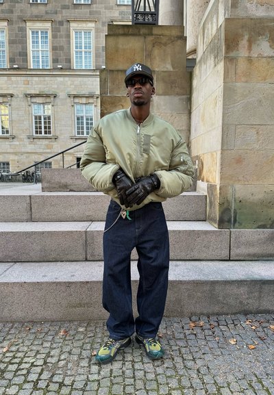 Chaqueta bomber verde aceituna con cremallera, guantes negros, pantalones vaqueros holgados azul oscuro y zapatillas verdes y negras, de pie en escalones de piedra.