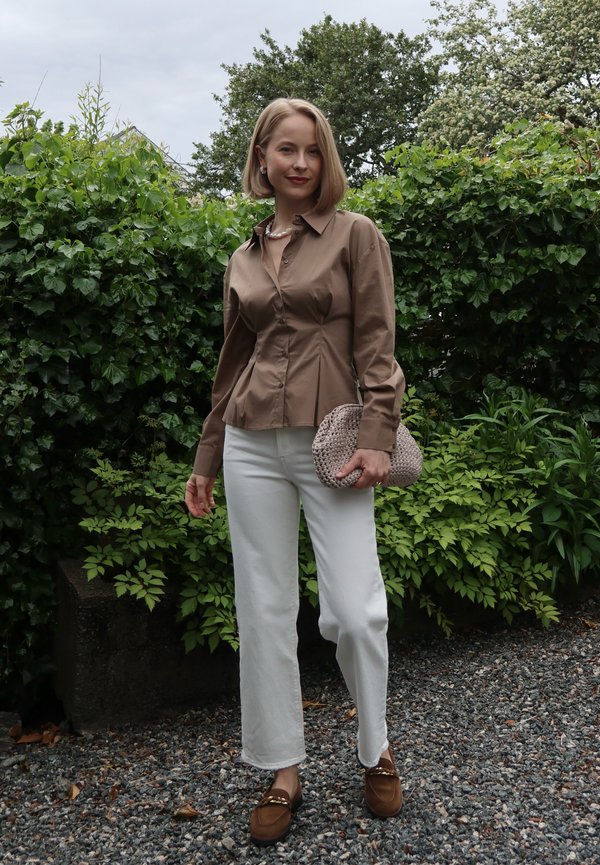 Femme debout à l'extérieur portant une chemise marron boutonnée, un pantalon blanc, des mocassins marron, un collier de perles, des boucles d'oreilles, et tenant une pochette tissée beige.