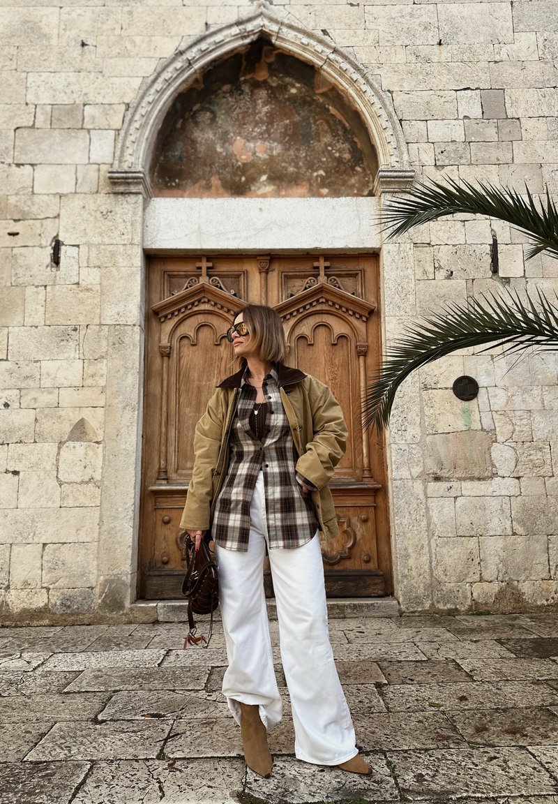 Femme portant des lunettes de soleil, une veste beige, une chemise à carreaux, un pantalon blanc et des bottes marron clair, debout devant des portes doubles en bois ornées avec un arc en pierre.