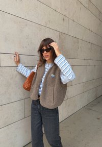 Brūns vilnas bezpiedurkņu vestes apģērbs virs zila un balta svītrainā krekla, kombinēts ar tumšiem džinsiem. Brūna austā rokassoma piešķir tekstūru. Fons ir siena.