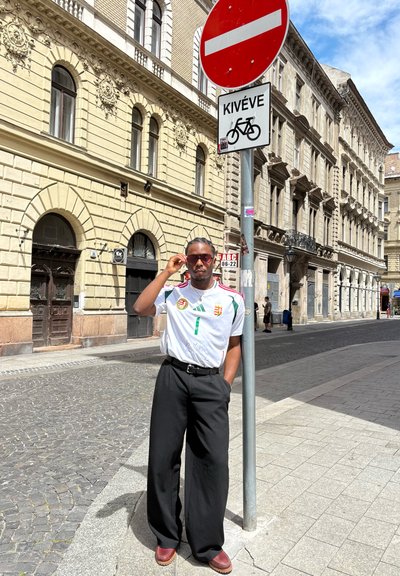 Camiseta deportiva blanca con detalles en verde y rojo, pantalones negros anchos y zapatos marrones. El cartel en el fondo indica un camino para bicicletas.