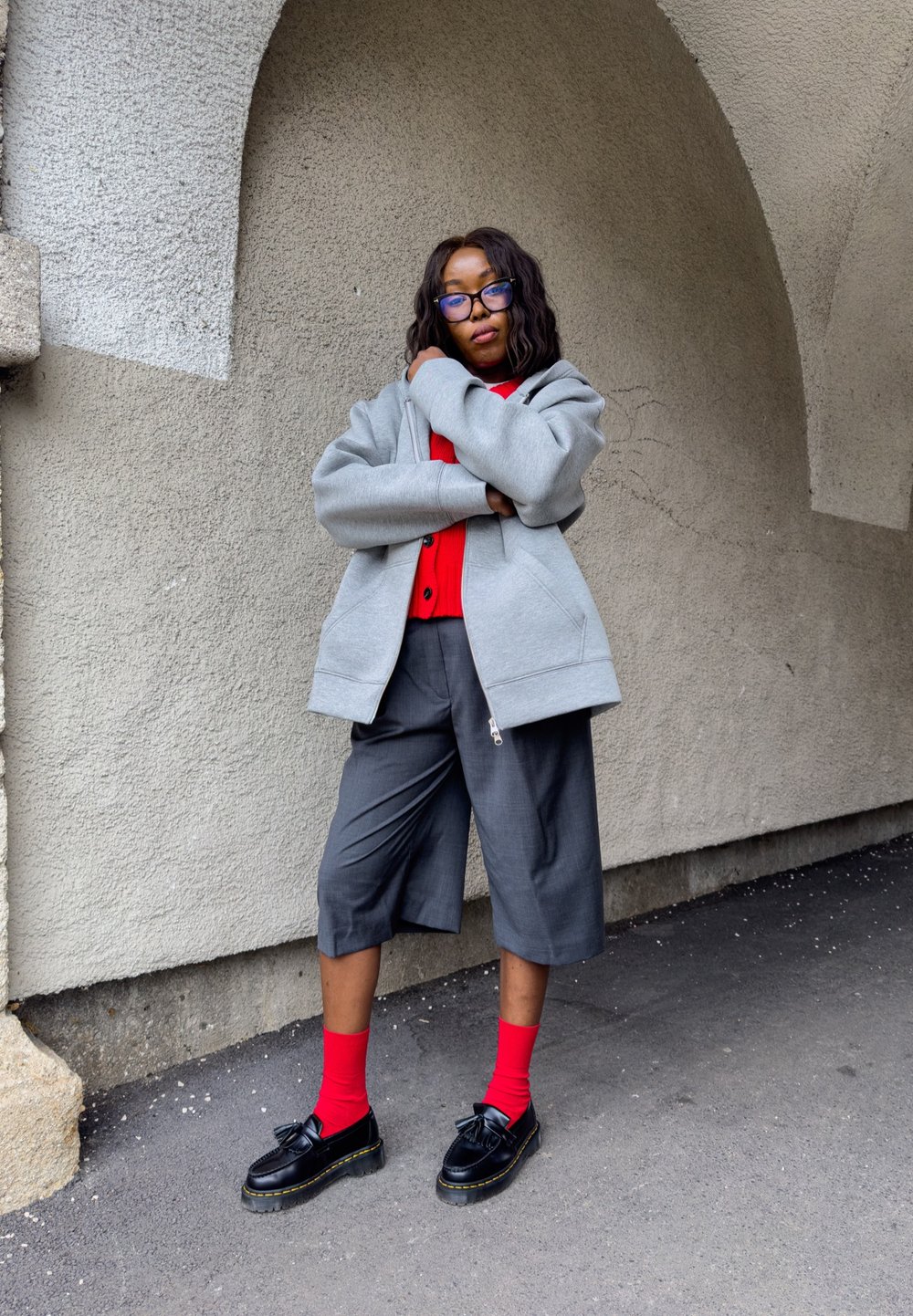 Felpa grigia con zip sopra una maglietta rossa, abbinata a pantaloni corti grigi, calze rosse e scarpe nere con suola spessa. Lo sfondo è una parete texturizzata.
