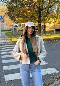 Jovem mulher vestindo um casaco bege e um chapéu, segurando um copo de café, parada numa faixa de pedestres com árvores de outono e edifícios ao fundo.