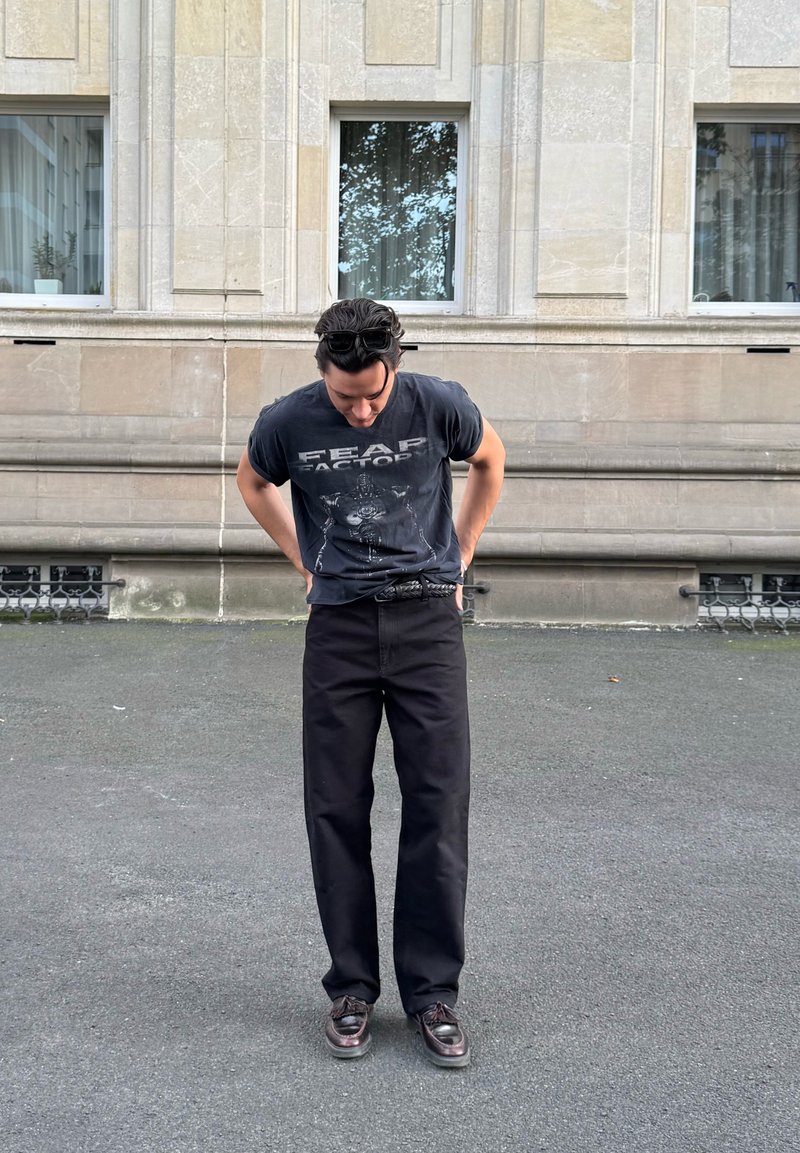 Un uomo con gli occhiali da sole sulla testa guarda in basso, indossando una maglietta nera "Fear Factory", pantaloni neri e scarpe di pelle scure, in piedi su una strada della città.
