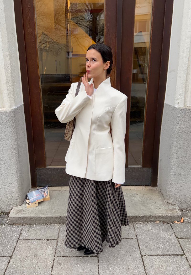 Femme en blazer blanc et longue jupe à carreaux debout sur le trottoir, tenant un sac à main, posant avec les doigts près des lèvres devant une porte en bois.
