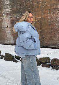 Femme aux longs cheveux blonds portant une veste matelassée bleu clair et un jean large, debout dans la neige près d'un mur marron avec des graffitis.
