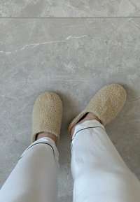 Pantofole in fleece beige con una superficie morbida e testurizzata, punta tonda e apertura sul retro, abbinate a pantaloni bianchi con piping neri.