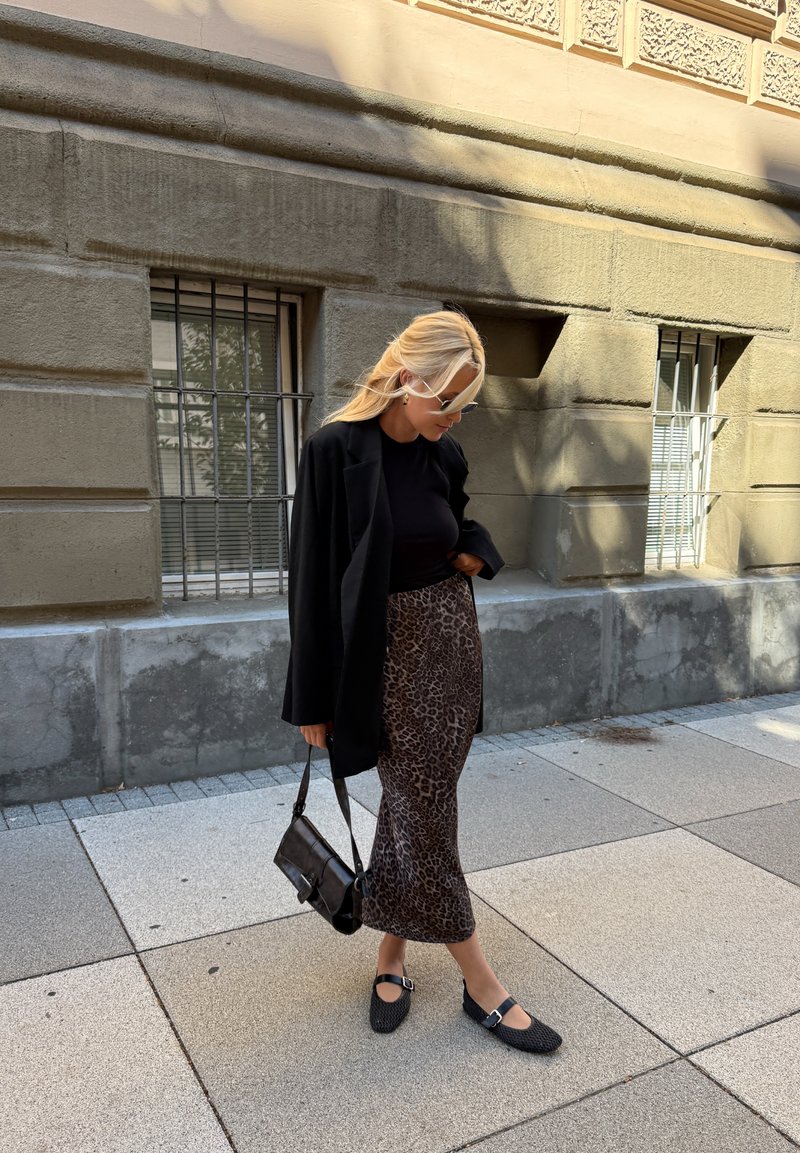 Femme portant une veste noire, un haut noir, une jupe à imprimé léopard, des chaussures noires et des lunettes de soleil marchant sur un trottoir à côté d'un bâtiment en pierre.
