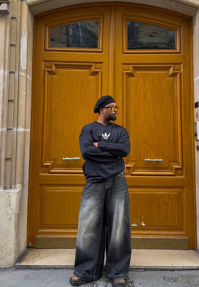 Hombre con boina negra, gafas de sol, sudadera negra de Adidas y pantalones vaqueros anchos descoloridos, de brazos cruzados frente a grandes puertas dobles de madera.