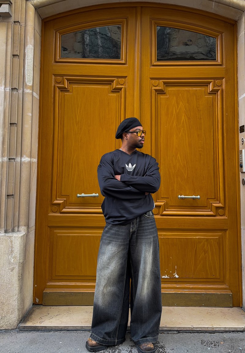 Mees, kes kannab musta baretti, päikeseprille, musta Adidas dressipluusi ja laiu, pleekinud teksapükse, seisab käed risti suures puitukse ees.