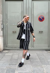 Μαύρη και γκρίζα ρίγα μπλούζα με κοντό μανίκι και σορτσάκι, συνδυασμένα με μαύρα παπούτσια και λευκές κάλτσες. Αξιοσημείωτα τατουάζ στα πόδια και ασημένια αξεσουάρ.
