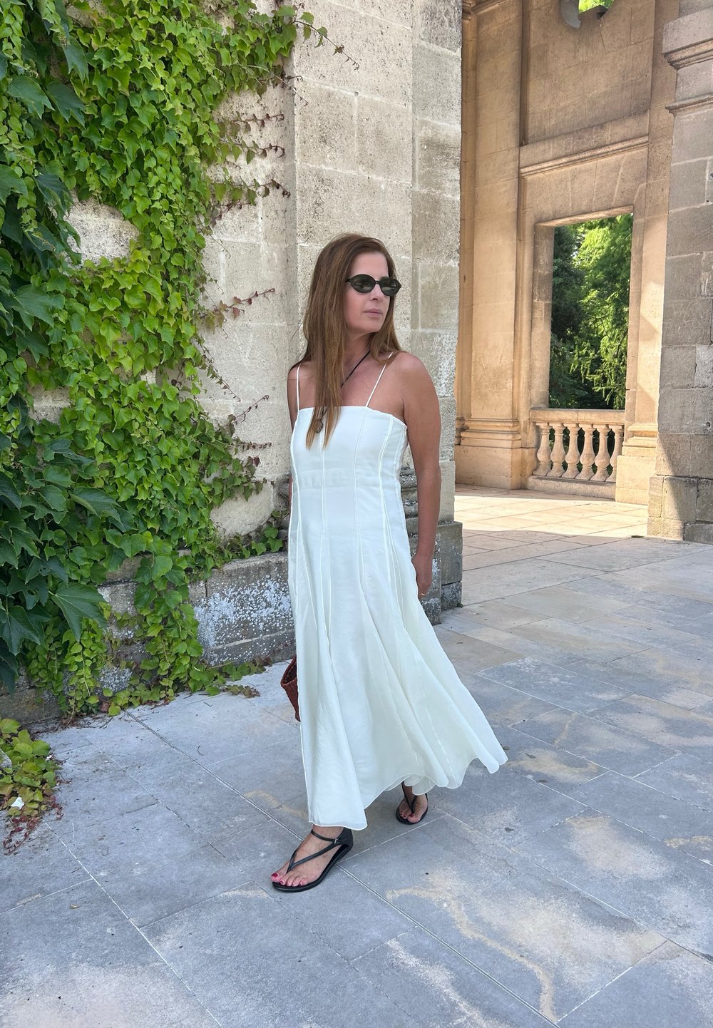 Weißes ärmelloses Maxikleid mit einem taillierten Oberteil und fließendem Rock, kombiniert mit schwarzen Flip-Flops. Im Hintergrund grüne Efeu und steinerne Architektur.
