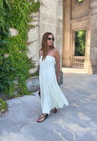 Weißes ärmelloses Maxikleid mit einem taillierten Oberteil und fließendem Rock, kombiniert mit schwarzen Flip-Flops. Im Hintergrund grüne Efeu und steinerne Architektur.