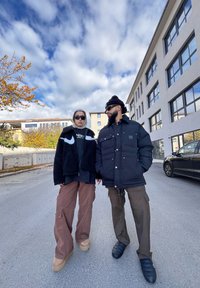 Δύο άτομα στέκονται έξω. Το ένα φοράει μια μαύρη, αφράτη ζακέτα με λευκές λεπτομέρειες και υπερμεγέθη καφέ παντελόνια. Το άλλο φοράει ένα μαύρο φουσκωτό μπουφάν και cargo παντελόνια.