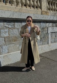 Manteau beige à double boutonnage, pantalon noir, chaussures claires et lunettes de soleil, adossé à un mur en pierre.