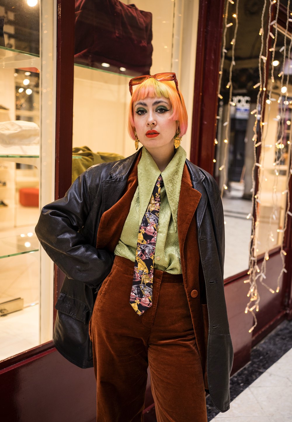 Completo in velluto a costine marrone con camicia verde e cravatta a fantasia. Giacca di pelle nera, occhiali da sole arancioni e capelli colorati. Vetrina sullo sfondo.