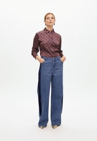 Camisa de botones marrón con estampado y cuello, combinada con unos vaqueros de pierna ancha en azul claro y zapatos metálicos de punta.