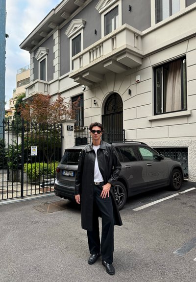 Abrigo de cuero negro sobre una camisa blanca, combinado con pantalones y zapatos negros. Edificio moderno y SUV oscuro en el fondo.