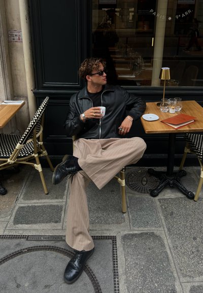 Chaqueta de cuero negra y camisa negra, pantalones de rayas en color beige, zapatos negros, sentado en una mesa con cristalería y un menú en un café al aire libre.