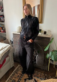 Črni pulover z zadrgo z vzorcem teksturiranega argyle, kombiniran z ohlapnimi temnimi hlačami. Okolje vključuje dekorativni kamin in rastline.