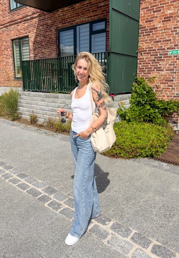 Ung kvinde med tatoveringer iført hvidt tanktop og blå jeans, der holder solbriller og en mulepose, stående på en brolagt gade ved en murstensbygning.