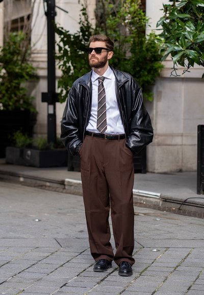 Hombre de pie en la acera de la ciudad, con gafas de sol, chaqueta de cuero negra, camiseta blanca, corbata a rayas, pantalones marrones y zapatos negros.