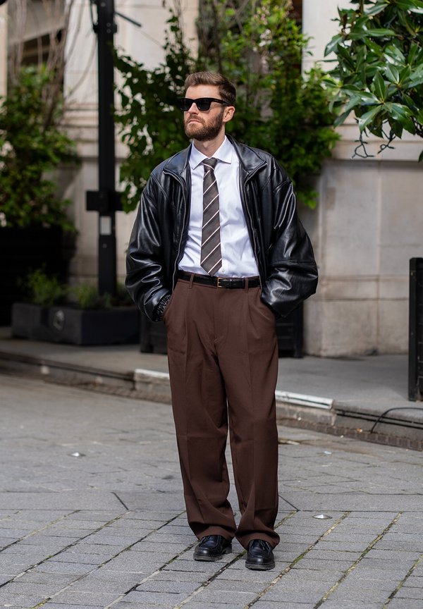 Man standing on a city pavement wearing sunglasses, black leather jacket, white shirt, striped tie, brown trousers, and black shoes.