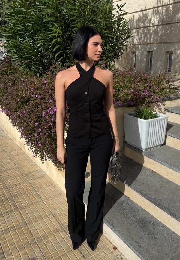 Ženska s črnim bob krojem las, oblečena v črni top z vrvico okoli vratu, črne hlače in koničaste čevlje, ki drži torbico z okrasnimi kroglicami blizu stopnic in rastlin.